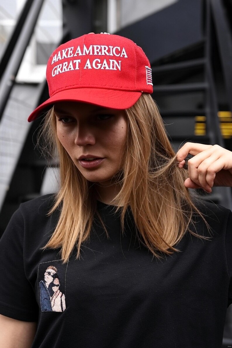 Puede incluir: Gorra de b&eacute;isbol roja con el texto blanco "MAKE AMERICA GRRAT AGAIN". La gorra tiene una visera curva y un peque&ntilde;o dise&ntilde;o de la bandera estadounidense en el lateral. La persona lleva una camiseta negra con un peque&ntilde;o parche bordado.