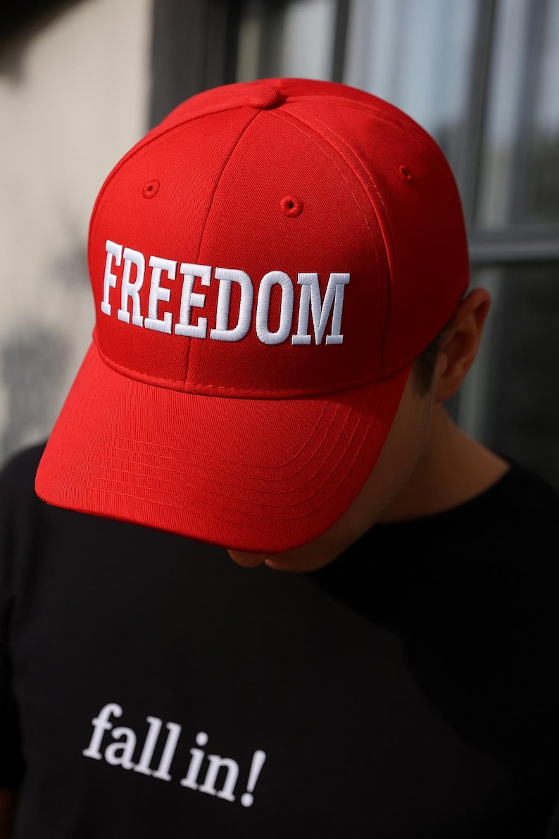 Puede incluir: Gorra de b&eacute;isbol roja brillante con la palabra "FREEDOM" bordada en blanco en la parte delantera. La gorra tiene una visera curva y un dise&ntilde;o estructurado. La persona lleva una camiseta negra con la frase "fall in!"