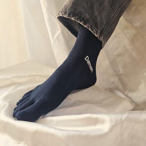 May include: Navy blue toe socks with the word "Damon" in white script. The socks are ribbed and cover the foot and toes separately. The person is wearing gray denim jeans.