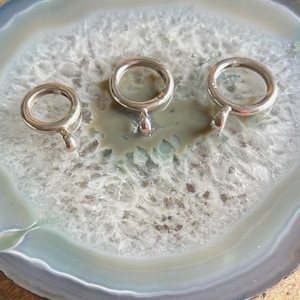 May include: Three silver curtain rings with a teardrop-shaped pendant are displayed on a gray and white agate slice. The rings vary slightly in size, showcasing a simple, elegant design. The agate slice has concentric bands of gray, white, and blue.