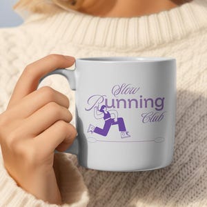 Peut inclure: Mug en céramique blanche avec un motif violet indiquant "Slow Running Club". Le motif représente une personne en train de courir. Le mug est tenu par une personne portant un pull blanc.