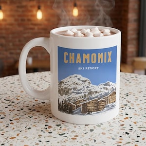 May include: White ceramic mug filled with hot chocolate and marshmallows. The mug displays a blue rectangle with the words "CHAMONIX SKI RESORT" and a mountain scene. Steam rises from the mug, suggesting a warm drink.