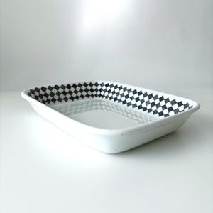 May include: A rectangular white ceramic baking dish featuring a black and white checkered pattern along the interior rim. The dish has a smooth, glossy finish and a shallow, open design with slightly sloped sides, suitable for oven use.