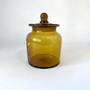 May include: A translucent amber-colored glass jar with a wide cylindrical body and a matching glass lid featuring a small rounded knob handle on top. The container has a smooth finish and a clear, hollow interior visible through the glass walls.