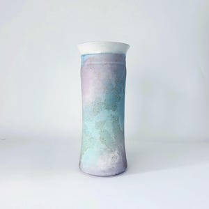 May include: A cylindrical ceramic vase featuring a smooth white rim and a mottled, watercolor-inspired glaze in shades of soft lavender, pale blue, and mint green. The surface texture appears slightly speckled with subtle, earthy brown flecks.