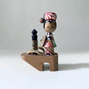 May include: A wooden Japanese doll with a red and white patterned hat and kimono. The doll stands on a wooden platform with a black cylindrical object and a rope. The doll has a serene expression and is a traditional craft.