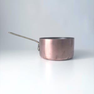 May include: A copper saucepan with a long, silver-colored handle. The pot has a rounded shape and a shiny, reddish-brown exterior. The handle is attached to the side of the pot. Kitchenware.