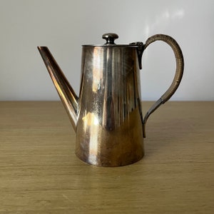 May include: A silver teapot with a brown handle. The teapot has a rounded body and a spout on the side. The handle is made of woven material.