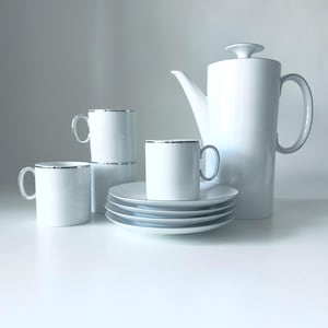 May include: A white porcelain coffee set with silver trim. The set includes a tall coffee pot, four mugs, and several saucers. The mugs and saucers are stacked. The set is displayed on a white surface.
