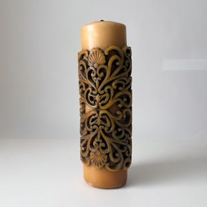 May include: A tall, brown pillar candle with an intricate, carved design. The candle is decorated with a repeating pattern of swirls and leaves.