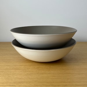 May include: Two stacked ceramic bowls. The bowls are off-white on the outside and gray on the inside. The rim of each bowl is gray. The bowls are sitting on a light brown wooden surface.