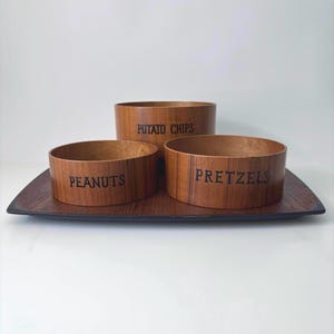 May include: A wooden serving set with three bowls labeled "Potato Chips", "Peanuts", and "Pretzels". The bowls are arranged on a dark wooden tray. The bowls and tray have a natural wood grain finish.