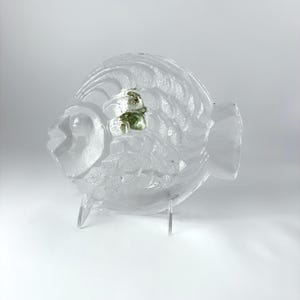 May include: A clear glass fish-shaped decorative plate featuring a textured scale pattern and a small, raised green glass accent near the center. The piece is supported by a transparent acrylic stand, showcasing its translucent form.