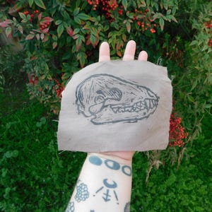 May include: A piece of tan fabric with a black linocut print of an animal skull. The fabric is held up against a backdrop of green grass and red berries.