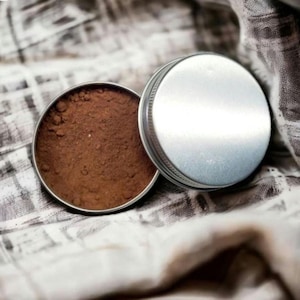 May include: Open, round, silver-colored tin with a matching lid. The tin contains a fine, brown powder. The lid is slightly ajar, revealing the product. The background is a neutral-toned, textured fabric.