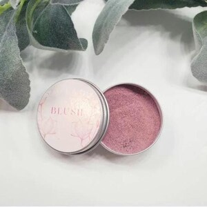May include: A small, round tin with a pink blush powder inside. The tin has a white label that says "BLUSH" in pink lettering.