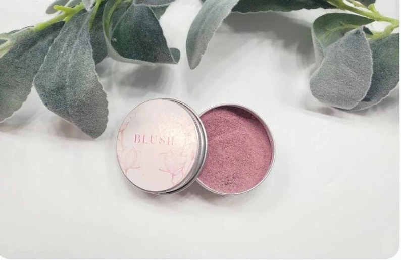 May include: A small, round tin with a pink blush powder inside. The tin has a white label that says "BLUSH" in pink lettering.