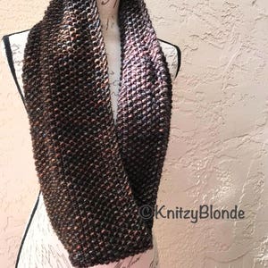 May include: A brown and black infinity scarf with a speckled pattern. The scarf is knitted and is displayed on a mannequin.