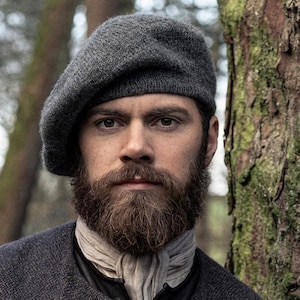 May include: A charcoal grey knit beret worn with a matching charcoal grey textured jacket featuring round buttons and a light beige neck scarf. The garment displays a fine, consistent weave pattern throughout the fabric of both the beret and jacket.