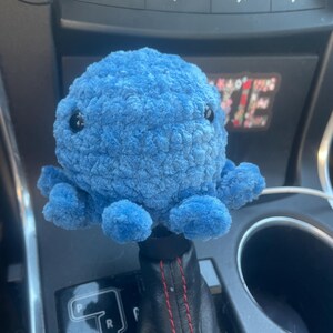 May include: A blue plush octopus gear shift knob with black eyes. The octopus is sitting on a black leather gear shift with red stitching. The gear shift has the letters P, R, N, S-D.