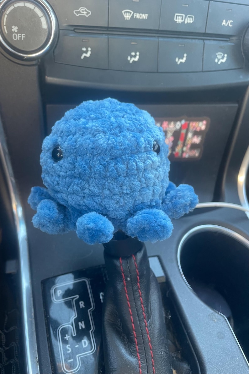 May include: A blue plush octopus gear shift knob with black eyes. The octopus is sitting on a black leather gear shift with red stitching. The gear shift has the letters P, R, N, S-D.