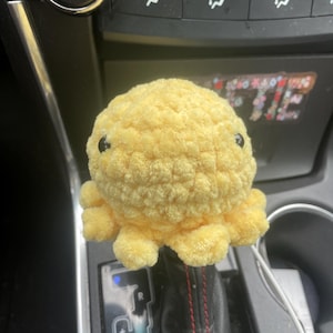 May include: A yellow crocheted octopus plush toy sits on a car's gear shift lever. The lever has a black leather boot with red stitching and a silver gear shift knob with the letters "R", "S-D", and "+" on it.