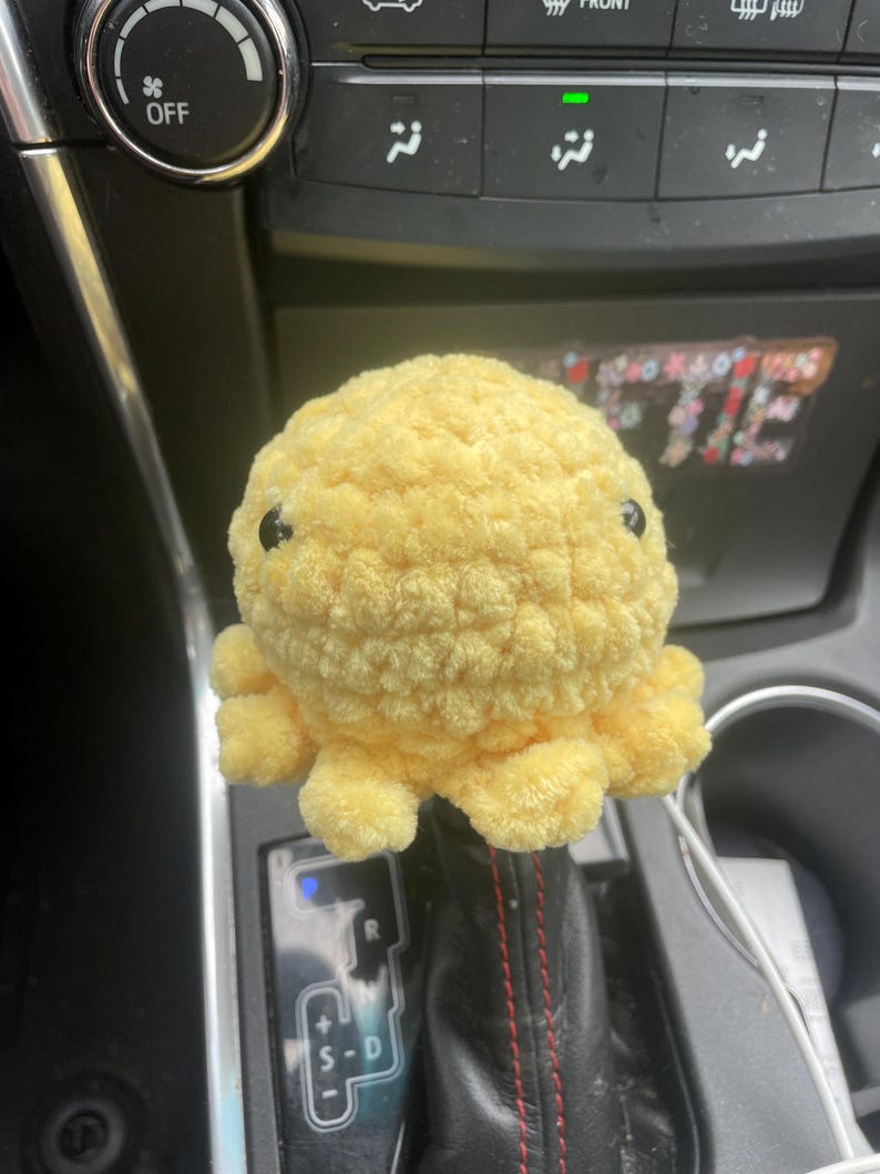 May include: A yellow crocheted octopus plush toy sits on a car's gear shift lever. The lever has a black leather boot with red stitching and a silver gear shift knob with the letters "R", "S-D", and "+" on it.