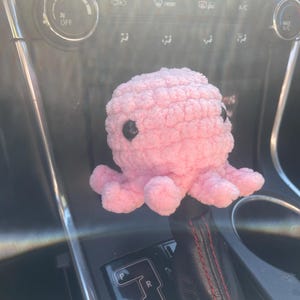 May include: A pink crocheted octopus with black eyes sits on a car's gear shift lever. The lever has a black leather cover with red stitching and a silver gear shift panel with the letters P, R, N, D, and +S-.