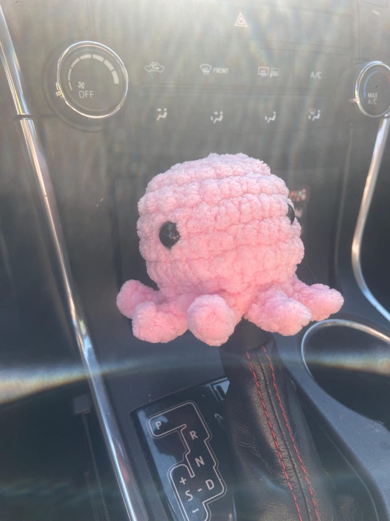 May include: A pink crocheted octopus with black eyes sits on a car's gear shift lever. The lever has a black leather cover with red stitching and a silver gear shift panel with the letters P, R, N, D, and +S-.