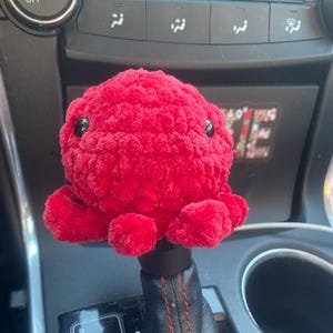 May include: A bright red crocheted octopus plush toy sits on a car's gear shift. The octopus has black eyes and a fuzzy texture.