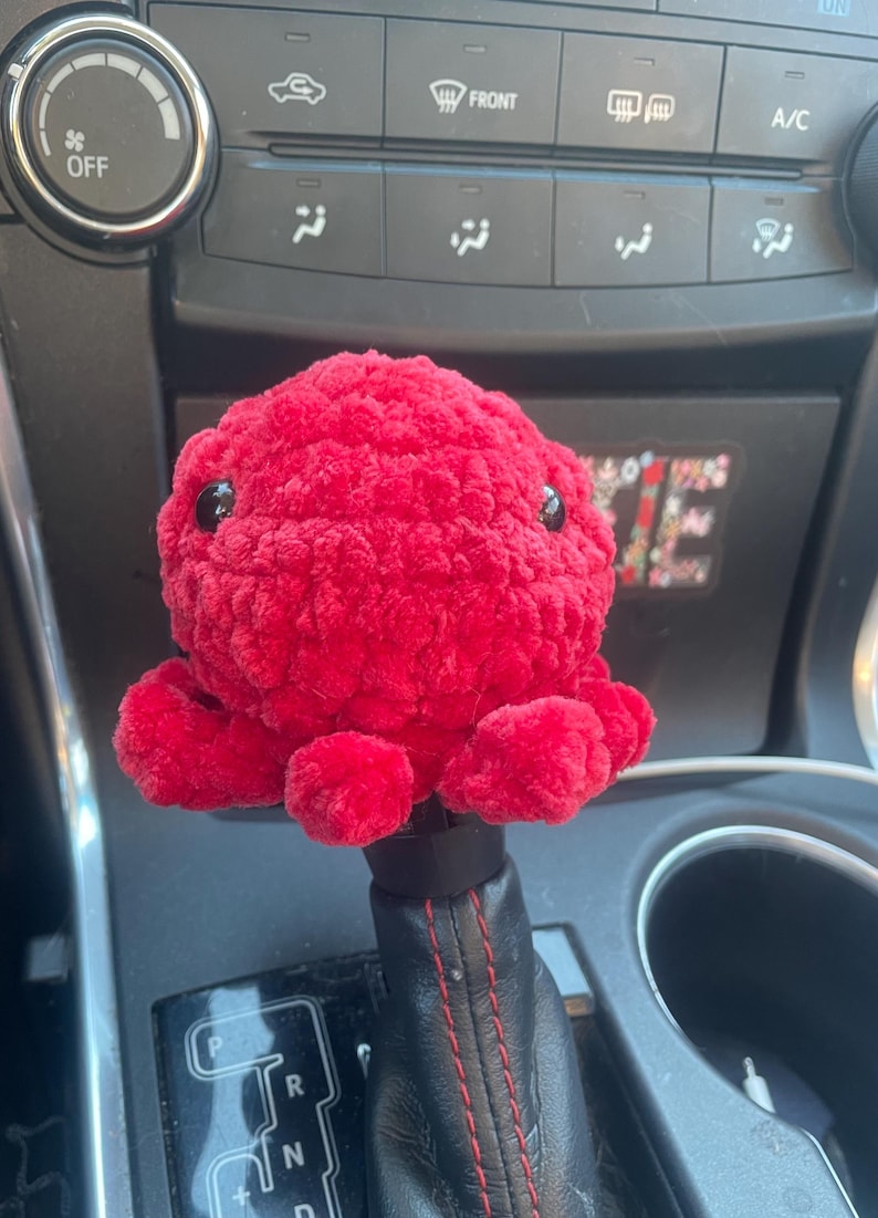 May include: A bright red crocheted octopus plush toy sits on a car's gear shift. The octopus has black eyes and a fuzzy texture.