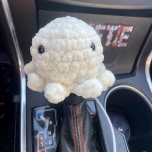 May include: White crocheted octopus car gear shift knob. The octopus has black eyes and is sitting on a black leather gear shift. The gear shift has a red stitching detail and the letters R, N, D, and S are visible.