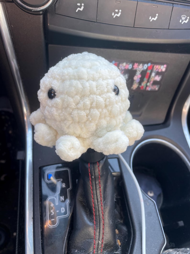 May include: White crocheted octopus car gear shift knob. The octopus has black eyes and is sitting on a black leather gear shift. The gear shift has a red stitching detail and the letters R, N, D, and S are visible.