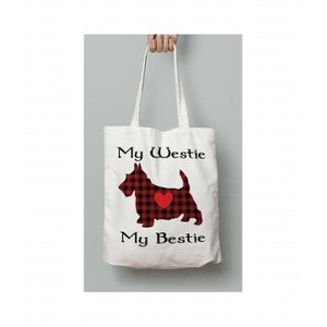 Puede incluir: Una bolsa de tela blanca con una silueta de West Highland White Terrier en tartán rojo y negro con un corazón rojo. El texto "My Westie My Bestie" está impreso en la bolsa.