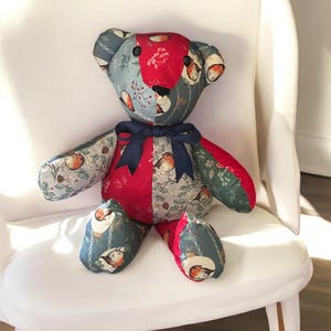 以下が含まれることがあります： ネイビーブルーのリボンが付いた手作りのパッチワークテディベア。クマの頭と手足は、花柄と鳥柄の赤、青、緑の生地で模様が施されています。体は赤とグレーです。クマは白い椅子に座っています。