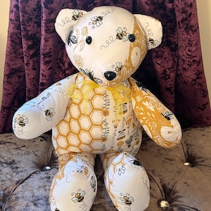 May include: A plush teddy bear with a white and yellow patchwork design featuring bees and honeycombs. The bear has black button eyes and a black nose.