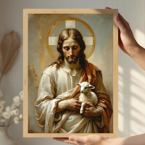 Könnte beinhalten: Ein gerahmtes religiöses Kunstwerk zeigt eine Figur mit langen braunen Haaren und Bart, die weiße und orangefarbene Gewänder trägt und ein weißes Lamm hält. Ein goldener Heiligenschein mit einem weißen Kreuz befindet sich hinter dem Kopf der Figur. Das Kunstwerk befindet sich in einem hellbraunen Holzrahmen.
