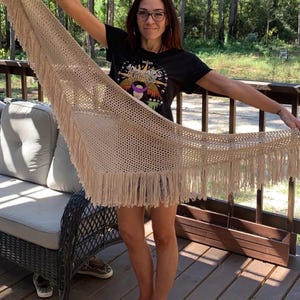Puede incluir: Un chal de crochet beige con flecos. El chal lo sostiene una persona que lleva una camiseta negra con el texto "Mysteries Purpose" impreso.