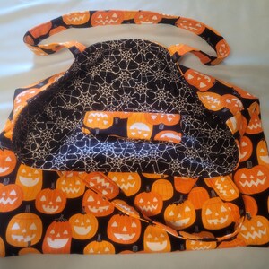 K&ouml;nnte beinhalten: Eine Halloween-Tragetasche mit einem K&uuml;rbismuster auf der Au&szlig;enseite und einem Spinnennetz-Design auf der Innenseite. Die Tasche zeigt orangefarbene K&uuml;rbisse mit l&auml;chelnden Gesichtern auf schwarzem Hintergrund. Die Griffe und der Besatz sind ebenfalls mit K&uuml;rbissen gemustert.