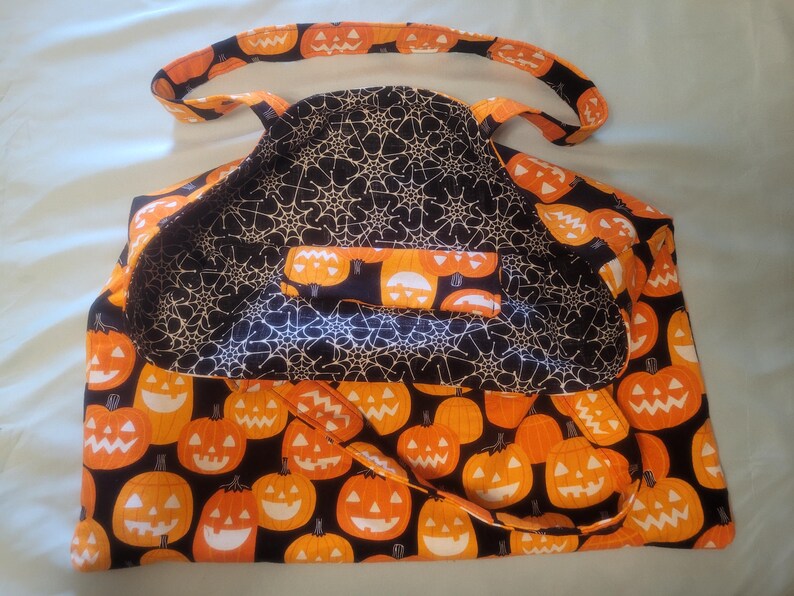 K&ouml;nnte beinhalten: Eine Halloween-Tragetasche mit einem K&uuml;rbismuster auf der Au&szlig;enseite und einem Spinnennetz-Design auf der Innenseite. Die Tasche zeigt orangefarbene K&uuml;rbisse mit l&auml;chelnden Gesichtern auf schwarzem Hintergrund. Die Griffe und der Besatz sind ebenfalls mit K&uuml;rbissen gemustert.
