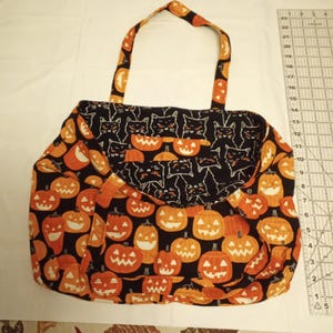 K&ouml;nnte beinhalten: Eine Halloween-Tragetasche mit schwarzem Hintergrund und orangefarbenen K&uuml;rbissen. Die Klappe zeigt schwarze Katzen. Die Tasche hat orangefarbene Griffe und ist aus Stoff. Die Tasche befindet sich auf einer wei&szlig;en Oberfl&auml;che.