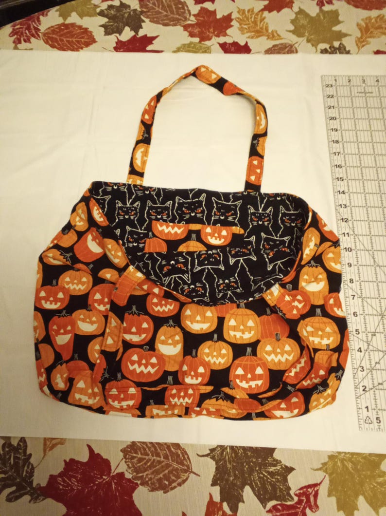 K&ouml;nnte beinhalten: Eine Halloween-Tragetasche mit schwarzem Hintergrund und orangefarbenen K&uuml;rbissen. Die Klappe zeigt schwarze Katzen. Die Tasche hat orangefarbene Griffe und ist aus Stoff. Die Tasche befindet sich auf einer wei&szlig;en Oberfl&auml;che.