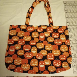 K&ouml;nnte beinhalten: Eine Halloween-Einkaufstasche mit schwarzem Hintergrund, bedeckt mit orangefarbenen K&uuml;rbissen. Die Tasche hat passende Griffe mit K&uuml;rbismuster. Die Tragetasche ist zum Tragen von Gegenst&auml;nden konzipiert.