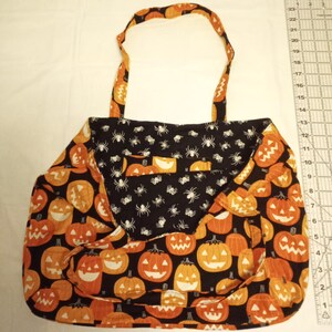 K&ouml;nnte beinhalten: Halloween-Tragetasche mit schwarzem Hintergrund und orangefarbenen K&uuml;rbissen. Die Tasche hat eine Klappe mit einem schwarzen Spinnennetz-Muster. Der Griff und die Verkleidung haben ein passendes K&uuml;rbismuster.