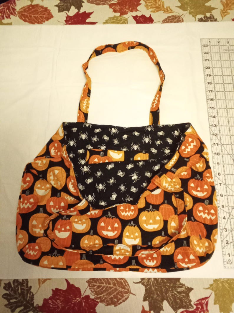 K&ouml;nnte beinhalten: Halloween-Tragetasche mit schwarzem Hintergrund und orangefarbenen K&uuml;rbissen. Die Tasche hat eine Klappe mit einem schwarzen Spinnennetz-Muster. Der Griff und die Verkleidung haben ein passendes K&uuml;rbismuster.
