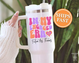 May include: White insulated tumbler with a handle, featuring the text "IN MY ENGAGED ERA" in pink, orange, and purple. Below, it reads "Future Mrs. Thomas" in black script. An orange circle with "SHIPS FAST" is visible.