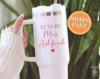 May include: White insulated tumbler with a handle, featuring the text "FUTURE Mrs. Ashford" in pink script, a small red heart, and a silver rim. The tumbler has a white lid and a straw. The design is for a wedding.