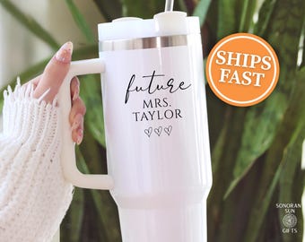 May include: White insulated tumbler with a handle, featuring the text "future MRS. TAYLOR" in black script, along with three heart symbols. The tumbler has a white lid and a silver rim. An orange circle with the words "SHIPS FAST" is visible.