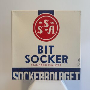 Könnte beinhalten: Eine Vintage-Illustration einer Zuckerdose mit dem Text "Bit Socker" und "Standard Kvalitet" in blauer Schrift. Die Dose zeigt außerdem einen roten Kreis mit den Buchstaben "S-S-A" in Weiß und einer blauen Krone über jedem Buchstaben. Der Text "Sockerfabriaget" steht in blauer Schrift am unteren Rand der Dose.