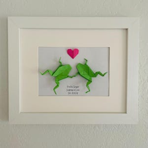 May include: A white framed wall art piece featuring two green origami frogs holding hands beneath a small pink paper heart. Below the frogs, the text reads: Fred & Ginger, Leaping in Love, Est. 01.01.01. The artwork is set against a plain white background.
